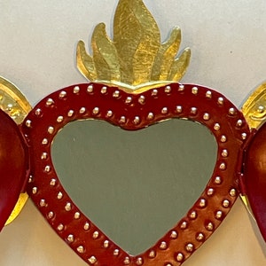 May include: A red heart-shaped mirror with a gold flame design on top. The mirror is surrounded by a border of small gold dots.