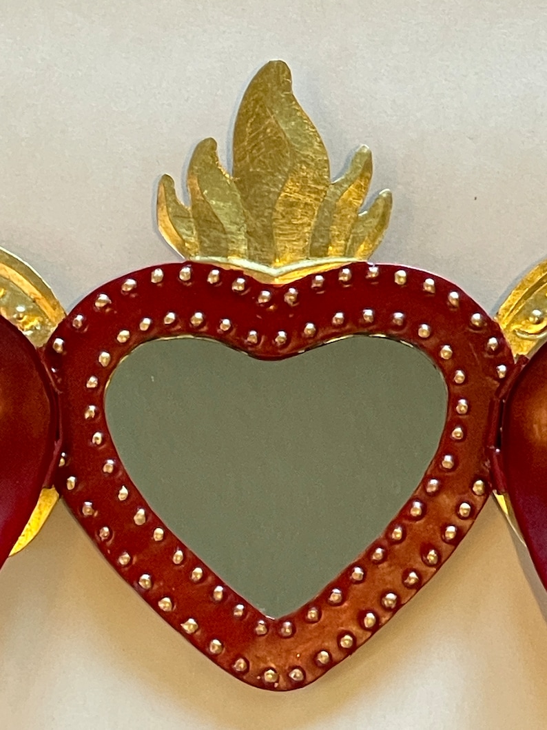 May include: A red heart-shaped mirror with a gold flame design on top. The mirror is surrounded by a border of small gold dots.