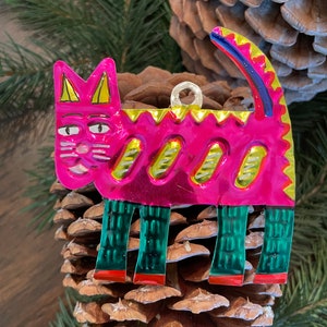 May include: A pink metal cat ornament with green and yellow details. The cat has a whimsical expression and is standing on a pine cone.