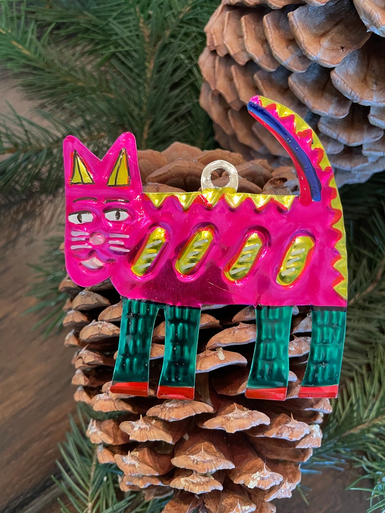 May include: A pink metal cat ornament with green and yellow details. The cat has a whimsical expression and is standing on a pine cone.