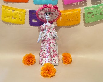 May include: A white paper mache skeleton doll wearing a pink and white floral dress and a pink hat with white lace trim. The doll is surrounded by three orange marigold flowers.