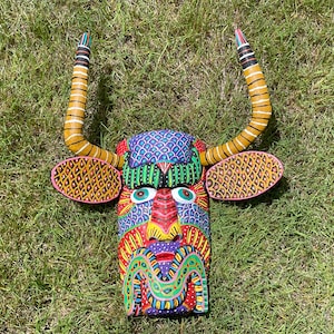 Puede incluir: Una máscara de madera pintada con colores vibrantes, con un diseño de toro. La máscara presenta un patrón multicolor con tonos azules, rojos, verdes y amarillos. Tiene cuernos grandes y curvos y formas parecidas a orejas. La máscara está sobre hierba verde.