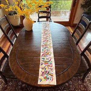 May include: A dark brown oval dining table with a colorful embroidered table runner. The runner features a floral and bird design in various colors. Surrounding the table are wooden chairs and a vase of yellow flowers.