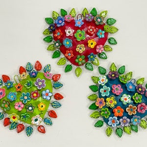 May include: Three heart-shaped metal ornaments with a floral design. Each heart is painted a different color: red, green, and blue. The flowers are painted in various colors, including pink, yellow, orange, and purple.