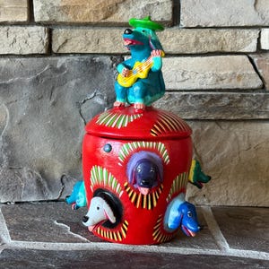 May include: A vibrant red ceramic jar with a lid, adorned with dog-themed designs. The jar features three dog faces in circular cutouts and a dog playing a guitar on top. The jar is painted with bright colors and decorative patterns.