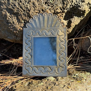 Może przedstawiać: Kwadratowa, metalowa rama z dekoracyjną, falistą krawędzią i wzorem promieni słonecznych u góry. Rama ma niebieskie wnętrze i jest umieszczona na naturalnym, skalistym tle. Rama ma wymiary około 13 cm kwadratowych.