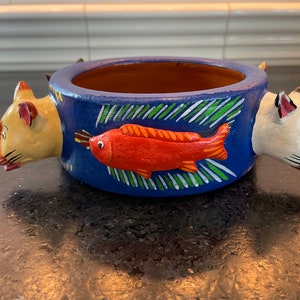 May include: A blue ceramic bowl with two cat heads on either side. The bowl has a red fish painted on the front with green and white accents.
