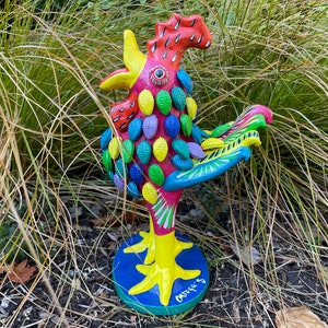May include: A colorful ceramic rooster figurine with a red body, yellow beak, blue tail feathers, and a green and purple patterned design. The rooster is standing on a blue and yellow base with the word "Oaxacas" written on it.
