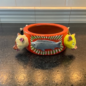 May include: A round orange ceramic planter with three animal faces around the rim. The faces are a white cat, a yellow cat, and a black cat. The planter has a green and white striped design with a gray fish painted on the front.