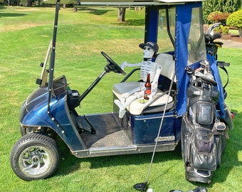 May include: A blue golf cart with a skeleton wearing a hat and holding a beer. The cart has a black golf bag with clubs and a golf club on the ground.