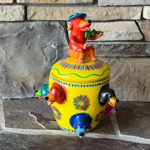 May include: A whimsical, hand-painted ceramic jar with a yellow base and lid. The jar features colorful dog heads emerging from the sides and a dog playing a violin on top. The jar is decorated with floral patterns and vibrant colors.