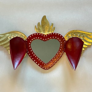 May include: A red heart with gold wings and a mirror in the center. The heart is decorated with gold studs and the wings have a gold leaf finish.