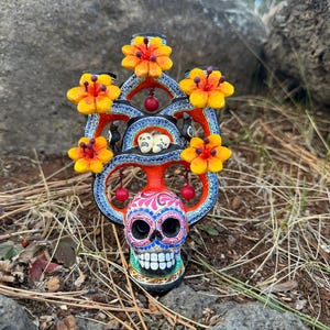 Peut inclure: Une sculpture de crâne du Jour des Morts avec un motif floral vibrant. Le crâne présente des motifs complexes et des couleurs vives, avec des fleurs jaunes et oranges ornant le dessus. Des accents rouges et un cadre bleu à motifs complètent le look festif.
