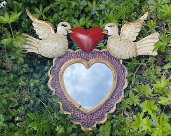 Puede incluir: Un espejo en forma de corazón con un marco dorado y un corazón rojo con dos palomas blancas a cada lado. El espejo está rodeado por un borde con estampado morado y dorado.