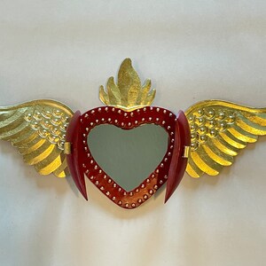 May include: A red heart with gold wings and a flame on top. The heart has a mirror inside and is decorated with small gold dots.