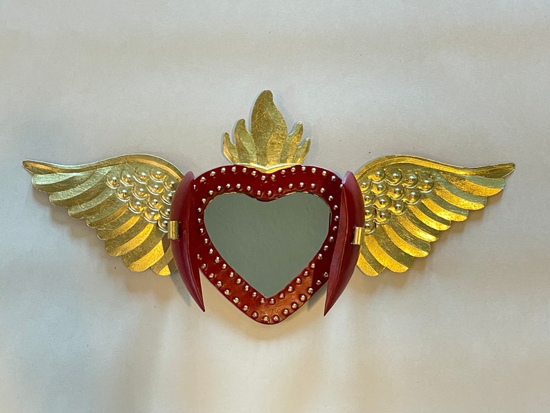 May include: A red heart with gold wings and a flame on top. The heart has a mirror inside and is decorated with small gold dots.