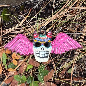 May include: A Day of the Dead-themed skull ornament with pink wings. The skull has a white base with black eye sockets and a wide, toothy grin. The wings are bright pink, and the skull is decorated with colorful patterns.