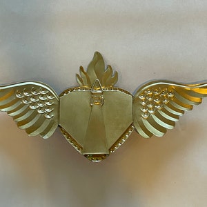 May include: Gold-colored metal picture frame shaped like a heart with wings. The frame has a textured surface and a small hook on the back for hanging.