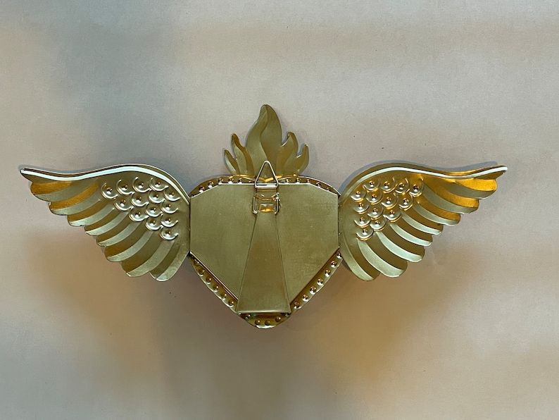 May include: Gold-colored metal picture frame shaped like a heart with wings. The frame has a textured surface and a small hook on the back for hanging.