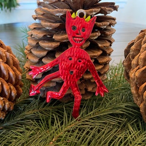 May include: A red metal devil-shaped ornament with yellow horns and a smiling face. The ornament is displayed on a pine cone, with other pine cones and evergreen branches in the background. A festive Christmas decoration.