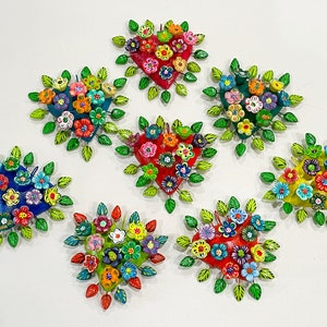 May include: Seven colorful ceramic hearts decorated with flowers and green leaves. Each heart is a different color, including red, blue, yellow, and green. The hearts are arranged in a circle on a white background.