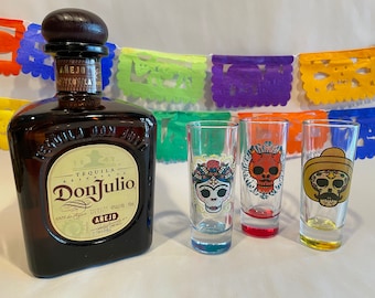 May include: A bottle of Don Julio Añejo tequila with a brown glass bottle and a gold label. Three shot glasses with colorful sugar skull designs are in front of the bottle. A colorful paper garland hangs in the background.