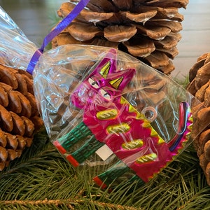 May include: A pink and green metal cat ornament with a shiny finish. The cat is in a clear plastic bag with a purple ribbon.