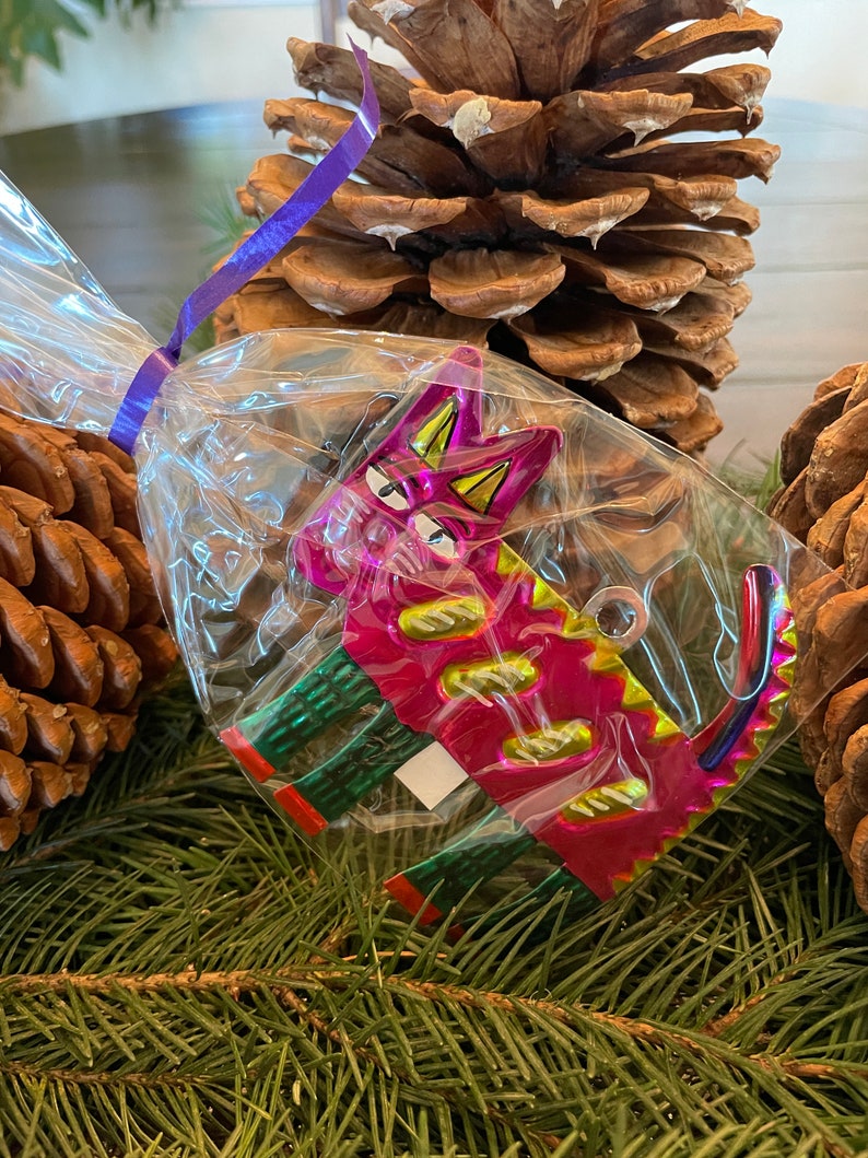 May include: A pink and green metal cat ornament with a shiny finish. The cat is in a clear plastic bag with a purple ribbon.