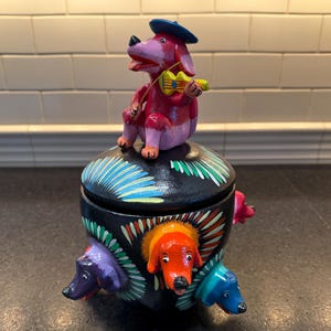May include: A black ceramic jar with a lid decorated with colorful painted dogs. The lid features a pink dog wearing a blue hat and playing a yellow violin.
