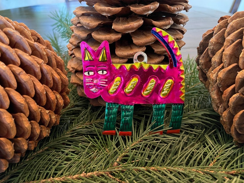 May include: A pink metal cat ornament with yellow and green details. The cat has a whimsical expression and is standing on green legs with red feet.