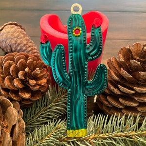 Könnte beinhalten: Ein grüner Weihnachtsbaumschmuck aus Metall in Form eines Kaktus mit roten und gelben Akzenten. Der Schmuck hängt an einem Kiefernzweig mit Tannenzapfen.