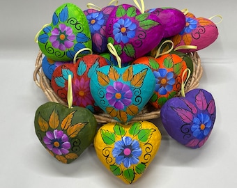 May include: A collection of colorful paper mache hearts with floral designs. Each heart is painted with vibrant colors and intricate details, including flowers, leaves, and swirls. The hearts are arranged in a basket, creating a festive and cheerful display.