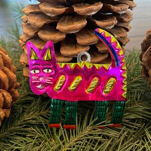 May include: A pink metal cat ornament with green and yellow details. The cat is standing on four legs and has a whimsical design.