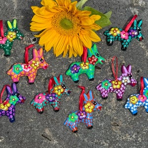 Op de afbeelding: Negen kleine ezelvormige ornamenten met levendige handgeschilderde bloemmotieven en stippen in diverse kleuren zoals groen, roze, blauw en paars, elk voorzien van een dun rood lintje om op te hangen, gerangschikt rondom een zonnebloem.