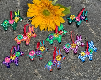 May include: Nine small donkey-shaped ornaments featuring vibrant hand-painted floral patterns and polka dots in various colors including green, pink, blue, and purple, each equipped with a thin red ribbon loop for hanging, arranged around a sunflower.