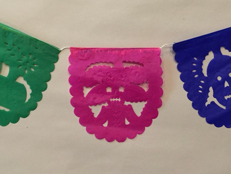 Puede incluir: Una cadena de tres banderines de papel con bordes festoneados. Los banderines son verdes, rosas y azules. Cada bander&iacute;n tiene un dise&ntilde;o recortado de una calavera.