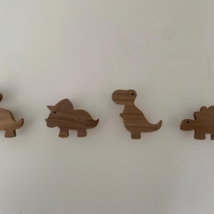 May include: Four wooden dinosaur-shaped hooks in various poses. The hooks are made of light brown wood and are all facing the same direction.