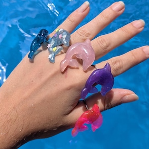 May include: A collection of five rings featuring dolphin-shaped ornaments in various colors including dark blue, glittery light blue with star accents, translucent pink, deep purple, and bright pink, each worn on a different finger of the hand.