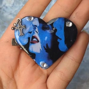 May include: Heart-shaped brooch with a blue and black image of two faces in profile, with red lips. The brooch has a silver-coloured cross charm and three clear rhinestones. The brooch is held in a hand.