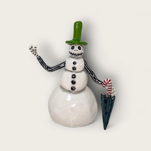 Puede incluir: Una caprichosa figura de muñeco de nieve con un sombrero de copa verde y brazos negros. El muñeco de nieve tiene una cara esquelética y detalles de botones negros. Sostiene un bastón de caramelo y un paraguas verde oscuro. Decoración navideña única.