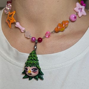 Puede incluir: Un collar de cuentas colorido con un colgante de un árbol verde con una cara. El collar incluye estrellas naranjas, huesos rosas y varias cuentas rosas y rojas. El colgante está detallado con una cara y una estrella.