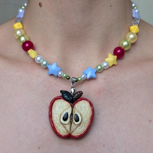 Puede incluir: Un collar de cuentas de colores con un dije de manzana roja. El collar tiene cuentas blancas, amarillas, rojas y azules, así como pequeños dijes de estrella azul. El dije de manzana es una representación realista de una manzana roja cortada por la mitad, con semillas visibles.