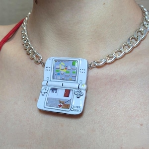 May include: A white pendant necklace featuring a miniature, painted replica of a handheld video game console. The game screen displays a colorful game scene. The pendant is attached to a chunky silver chain. The bottom of the console has the words "STAR SELECT".