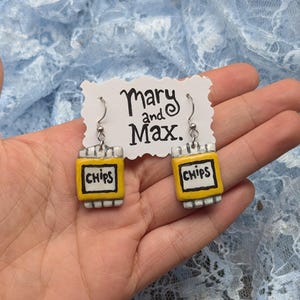 Puede incluir: Pendientes hechos a mano con miniaturas de bolsas de patatas fritas amarillas con la palabra "CHIPS" en un recuadro negro. Los pendientes están sujetos a ganchos plateados y a una etiqueta blanca con el texto "Mary and Max."