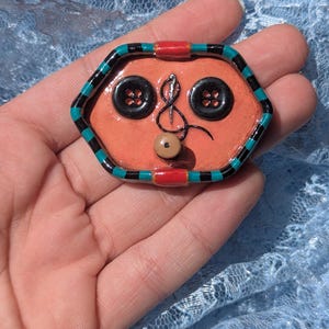 Puede incluir: Broche hexagonal naranja hecho a mano con ribete negro y verde azulado. El broche presenta dos botones negros, un diseño de aguja e hilo y una pequeña cuenta marrón. El broche se sostiene en una mano.