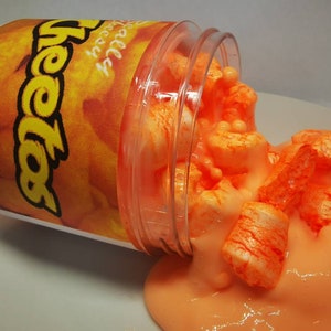 Puede incluir: Un recipiente de pl&aacute;stico transparente con una etiqueta naranja y amarilla que dice "Cheetos" est&aacute; derramando slime naranja con malvaviscos blancos.