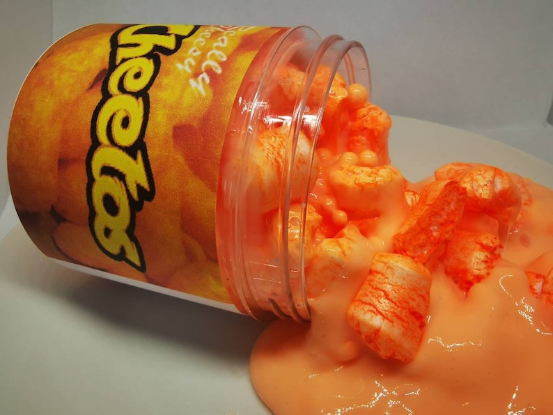 Puede incluir: Un recipiente de pl&aacute;stico transparente con una etiqueta naranja y amarilla que dice "Cheetos" est&aacute; derramando slime naranja con malvaviscos blancos.