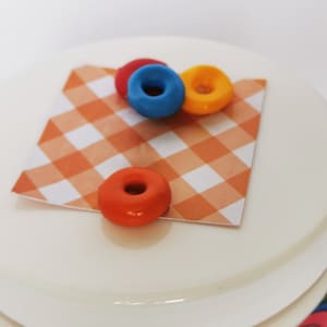 May include: A white ceramic jar with a red, white, and orange checkered fabric lid. Three small, colorful donuts are on top of the lid. The donuts are red, blue, and yellow.