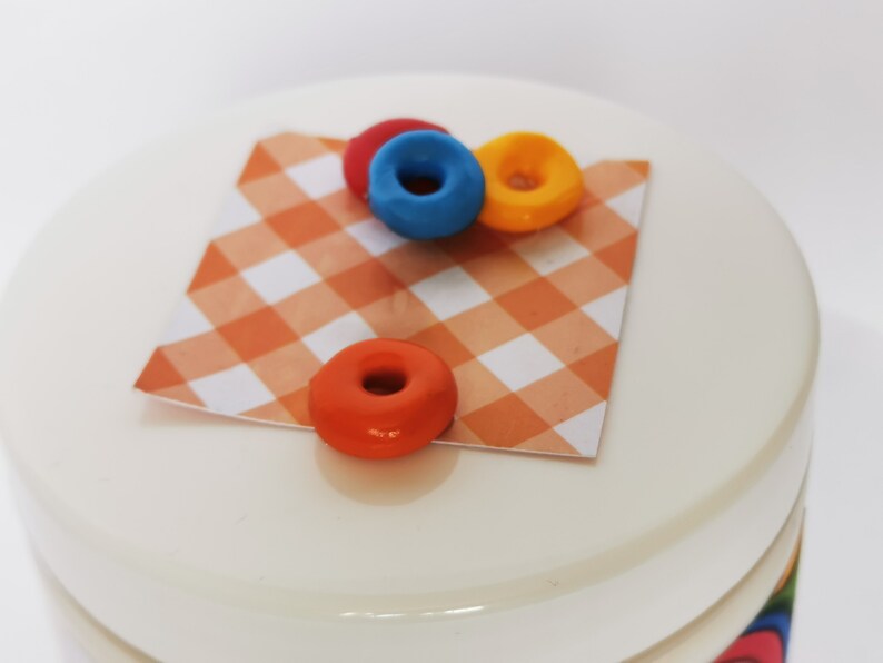 May include: A white ceramic jar with a red, white, and orange checkered fabric lid. Three small, colorful donuts are on top of the lid. The donuts are red, blue, and yellow.