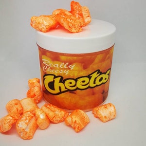Puede incluir: Snacks de Cheetos de color naranja en un frasco transparente con tapa blanca. El frasco tiene una etiqueta que dice "Really Cheesy Cheetos".